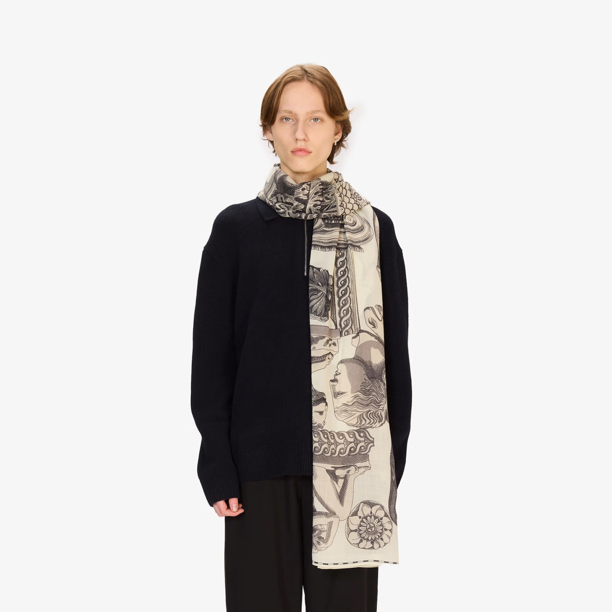 A model wearing a black knit zip-up sweater and a long cream-colored scarf with a black and gray graphic print, set against a plain light gray background.