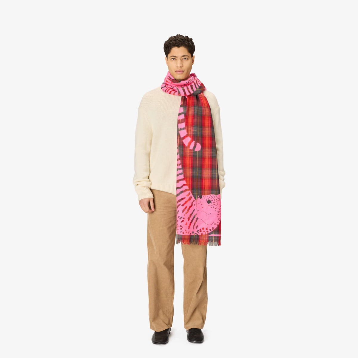 A model stands against a light gray studio background wearing a cream sweater, beige trousers, and a long scarf with a split design: one half is red and green plaid, and the other half is pink with a tiger motif.