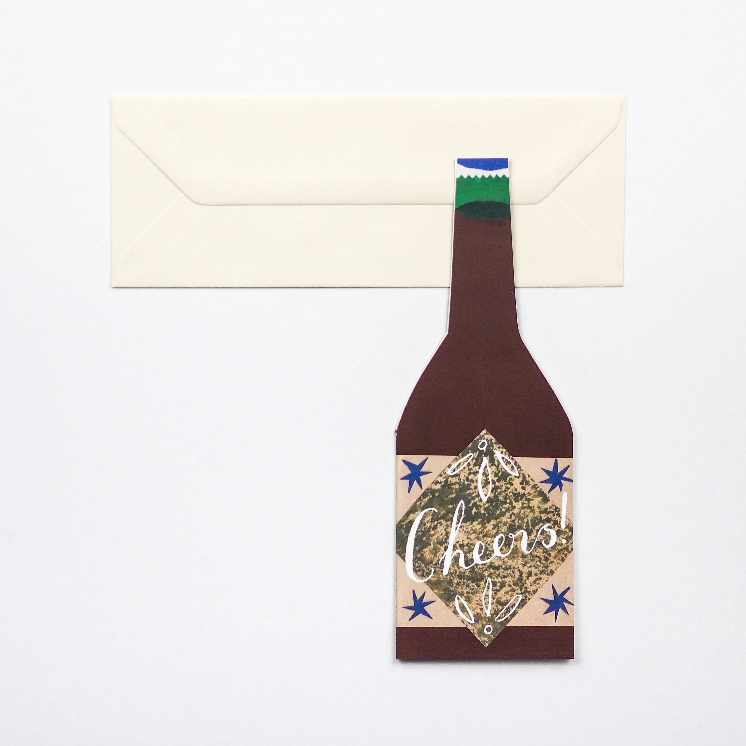 Paper beer bottle-shaped greeting card with "Cheers!" and stars; atop a cream envelope.