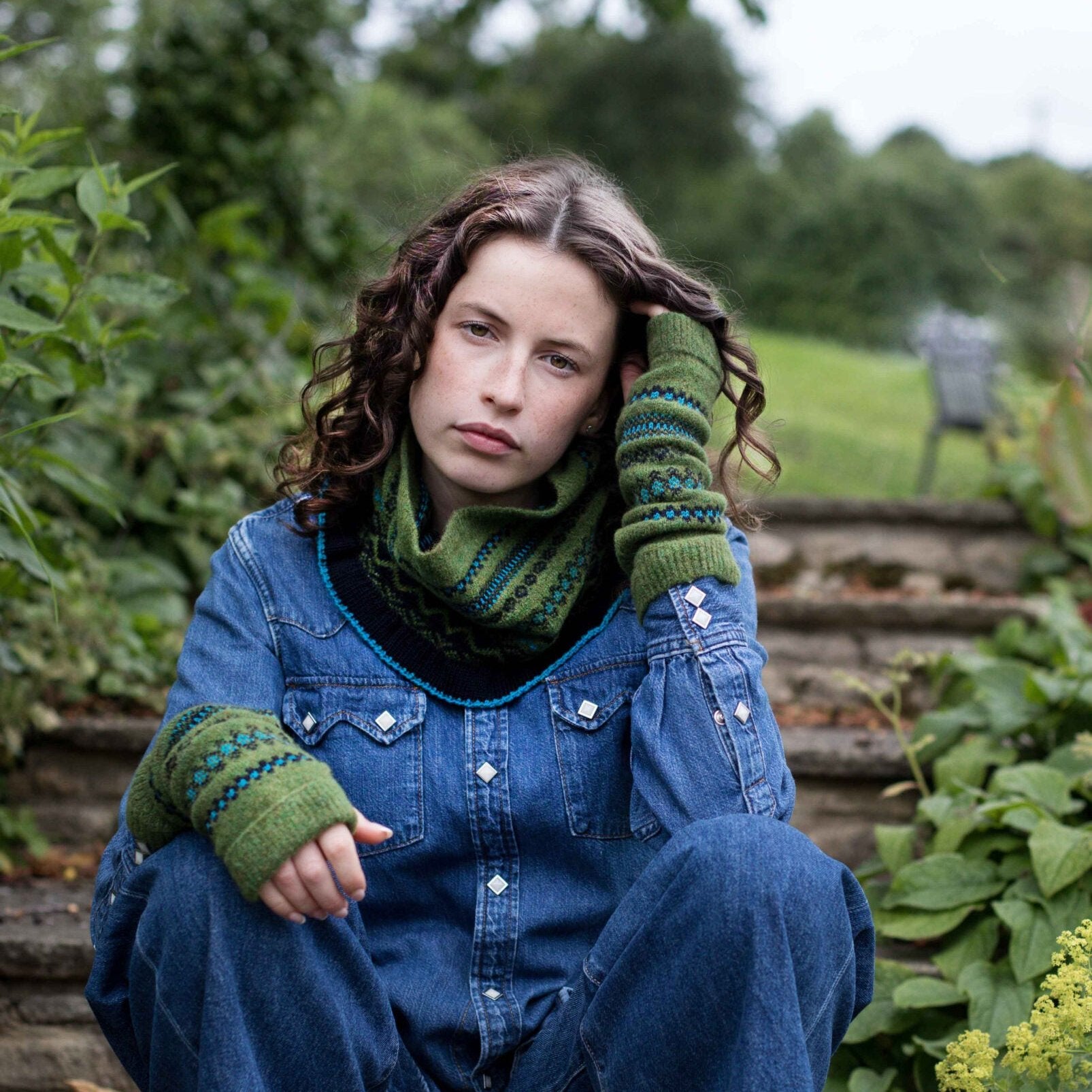 Green hand-knitted cowl and matching arm warmers with teal and black detailing.  The cowl is worn over a denim shirt.