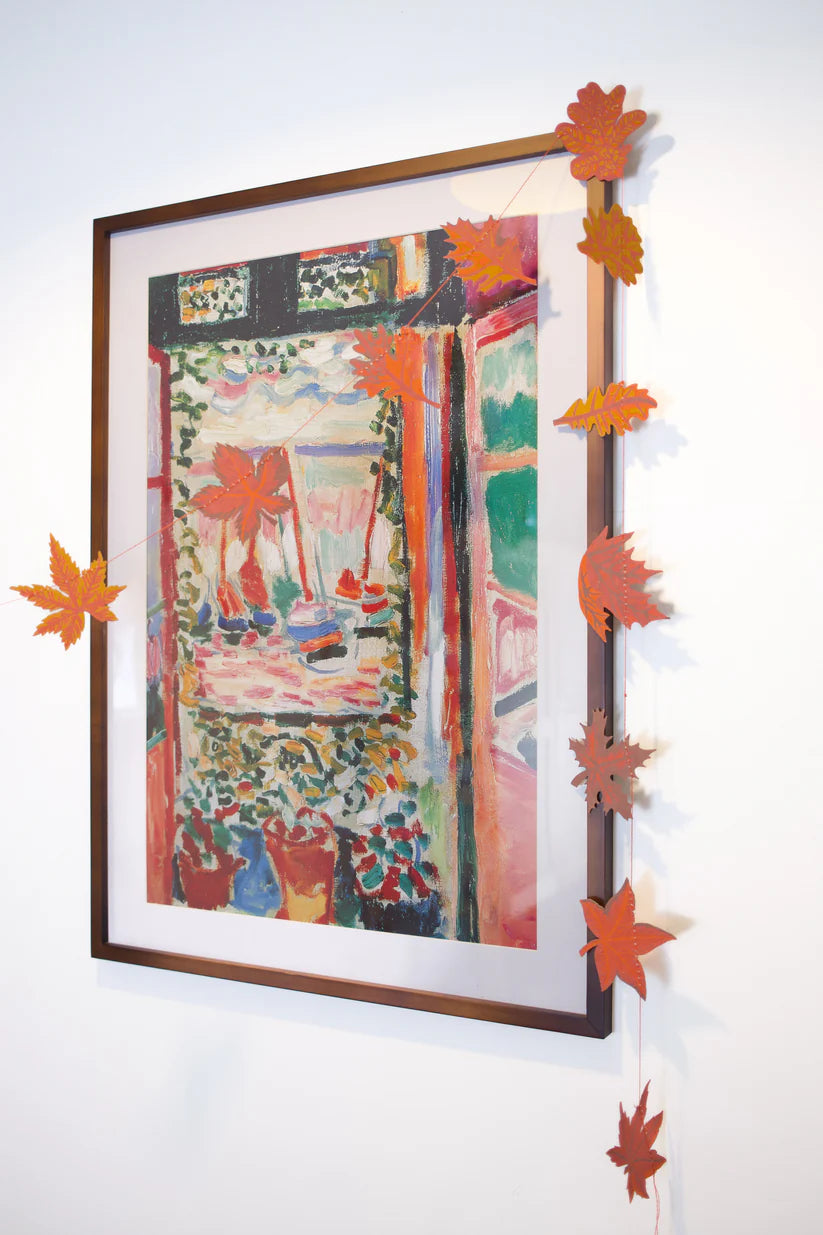 Framed painting depicting a vibrant indoor scene with potted plants and a window view, adorned with a fall leaf garland.