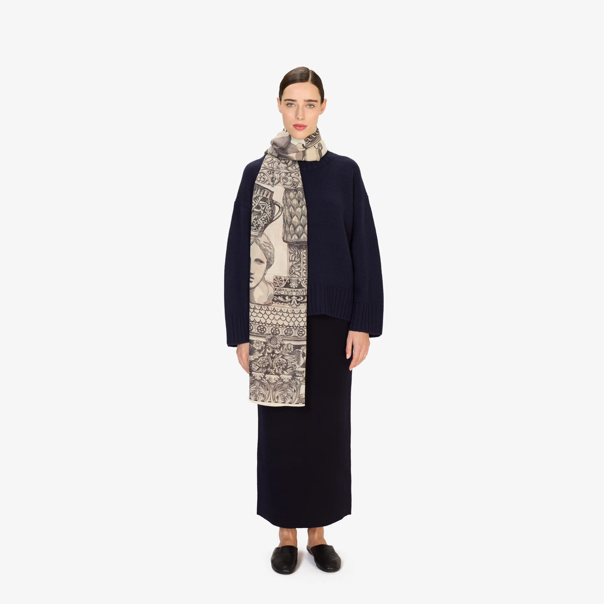 A model stands against a light gray studio background wearing a dark navy long-sleeve knit dress or sweater skirt, a beige and black patterned scarf, and black slip-on shoes.