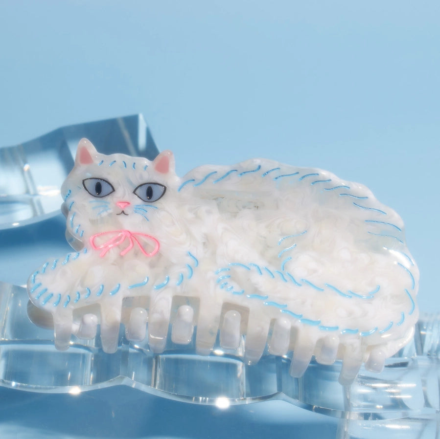 A white cat-shaped hair claw clip with blue stitched details, pink ears and nose, and blue eyes, resting on a clear acrylic stand against a light blue background.