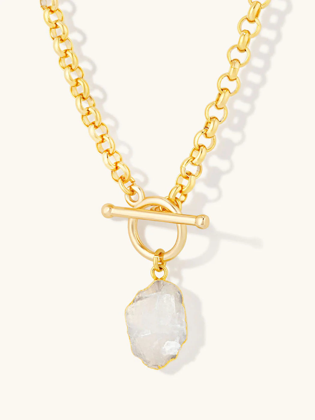 A gold chain necklace with a toggle clasp and a raw, irregular-cut clear gemstone pendant, shown on a light cream background.