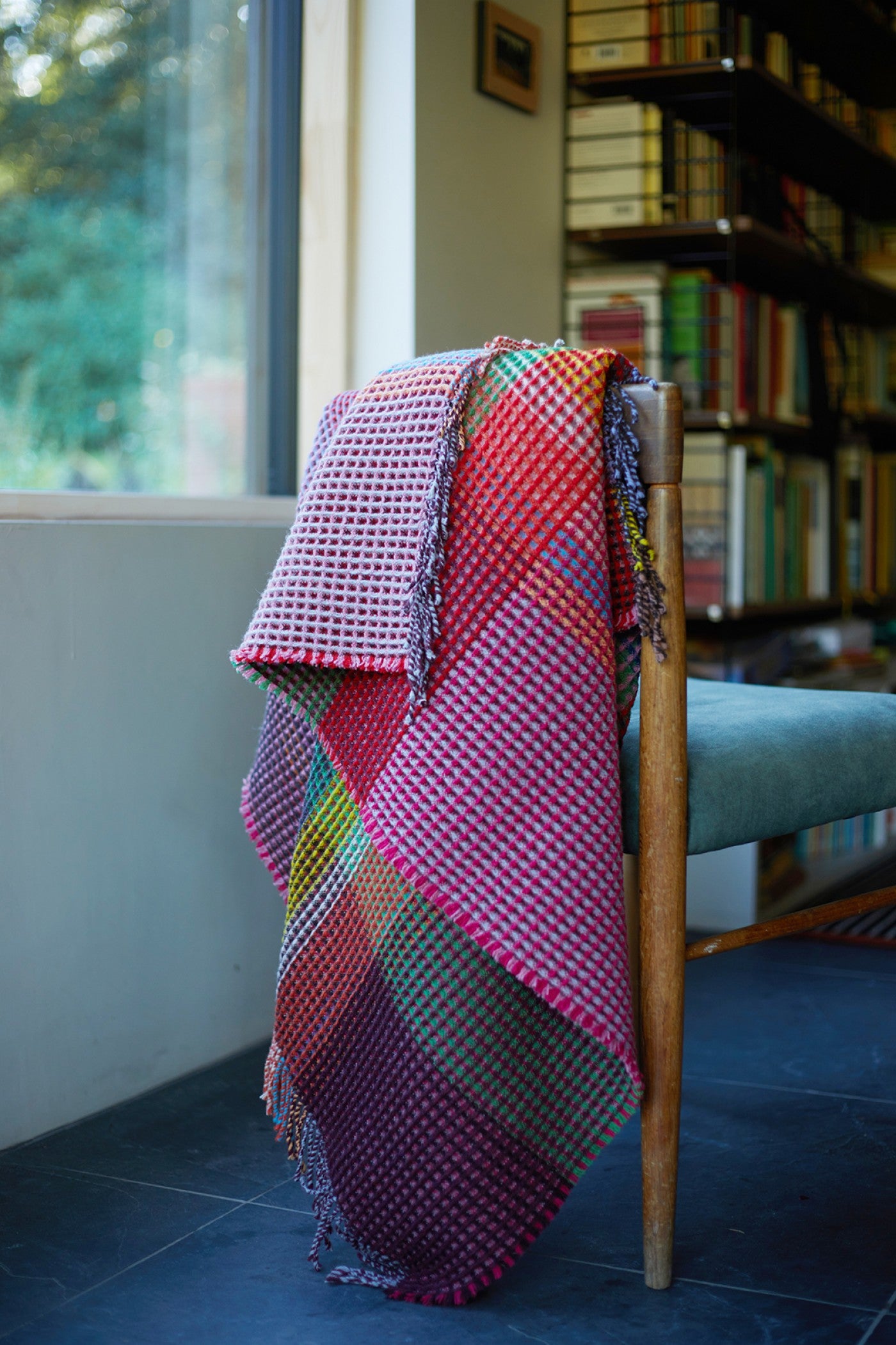 A colorful woven throw blanket with a pink, red, and purple plaid pattern draped over the back of a wooden chair in a home interior setting.