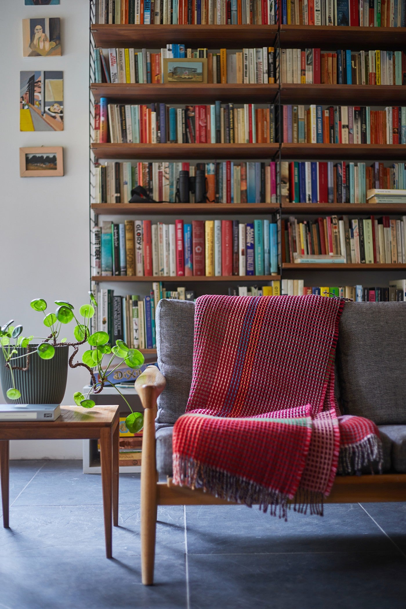 A cozy living room corner featuring a mid-century modern wooden armchair with a red and purple striped knit throw blanket draped over it, set against a large wall bookshelf filled with books.