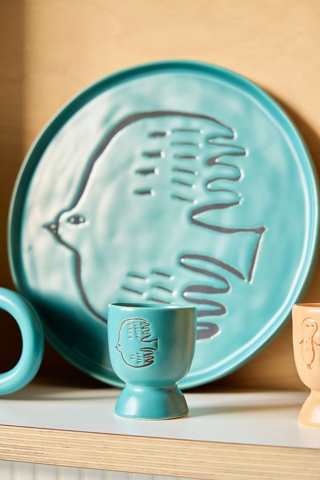 A turquoise ceramic egg cup with a debossed bird design and the text 'COOL TONY' in the center, displayed in front of a matching round turquoise serving platter on a light wood shelf.