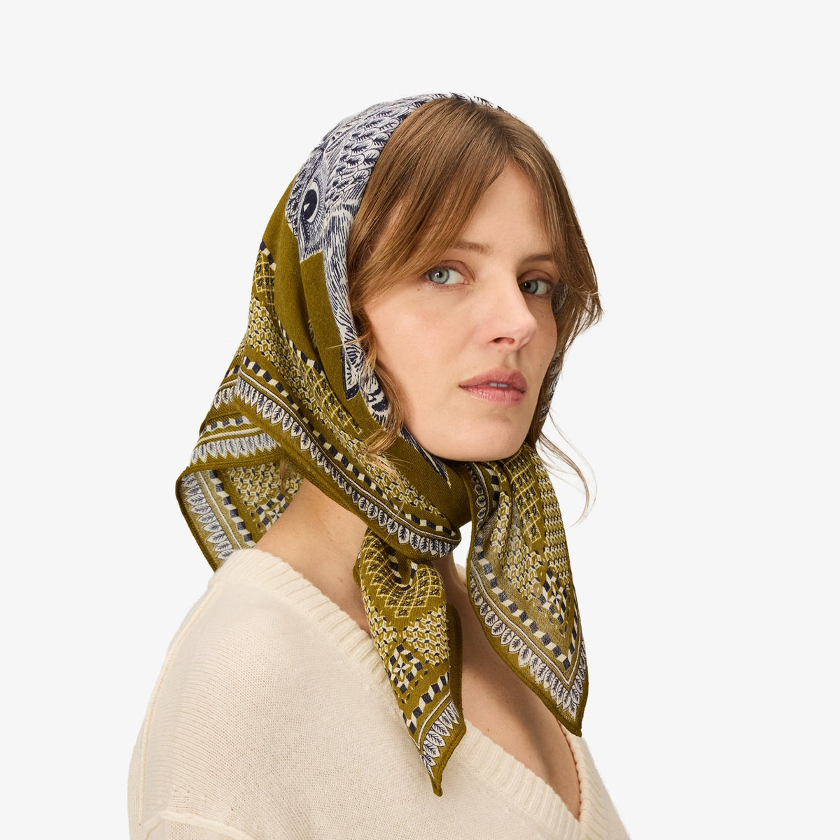 A woman wearing a square silk headscarf in olive green and white with a geometric tribal pattern, styled as a headband. She is wearing a cream-colored knit sweater against a solid light gray background.