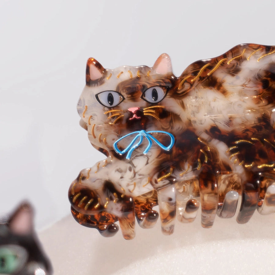 A close-up of a translucent claw hair clip shaped like a cat, with brown and cream marbled detailing, gold glitter accents, and a small blue bow on the face.