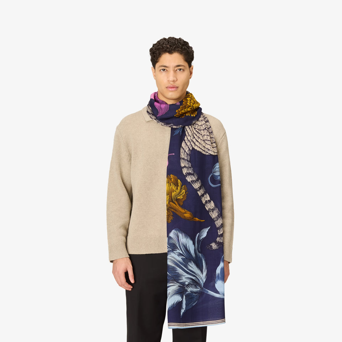 A model wearing a beige knit cardigan, black trousers, and a split-design scarf. One half of the scarf is a solid deep navy blue with a large light blue floral print, while the other half features a detailed illustration of a winged snake wrapped around a golden apple, set against a light gray background.
