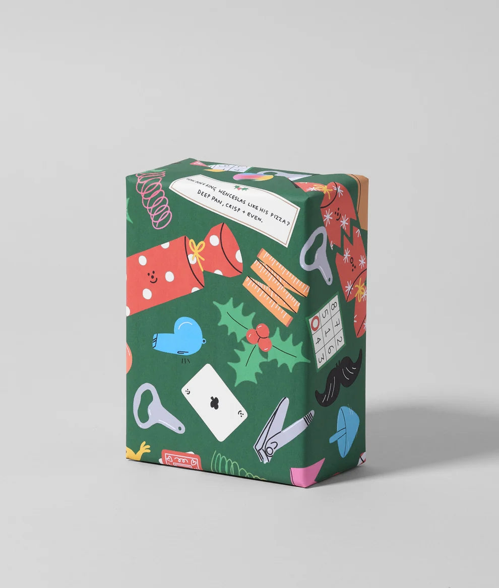 A square gift box wrapped in festive dark green paper with a playful pattern of holiday-themed illustrations, including a red polka-dot sweater, a red mug, a blue heart, holly leaves, and playing cards, set against a neutral light gray background.