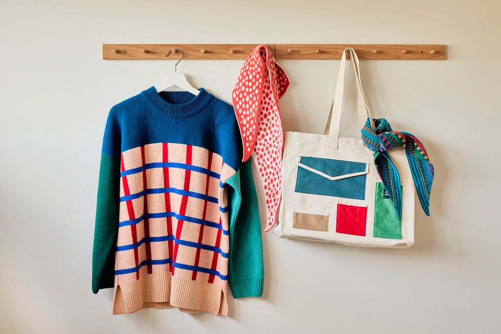 A colorful knit sweater hangs on a wooden wall rack alongside a red-and-white polka dot scarf and a cream canvas tote bag with patchwork pockets, all against a plain light wall.