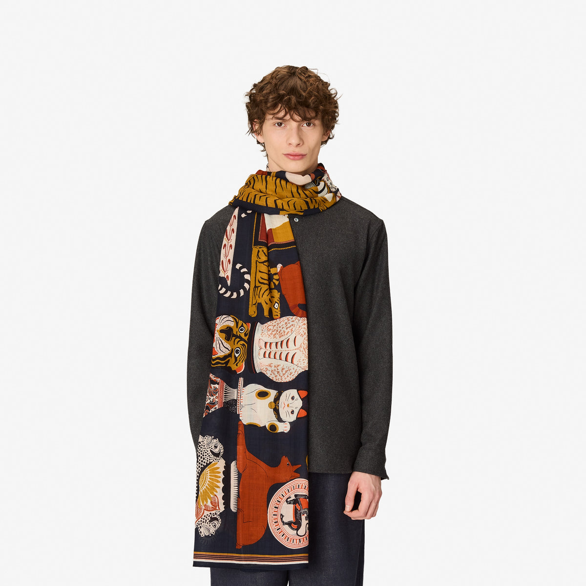 A model wearing a dark charcoal gray cardigan and a long, multi-colored patterned scarf featuring illustrations of Japanese foxes and cats in orange, cream, and navy blue.