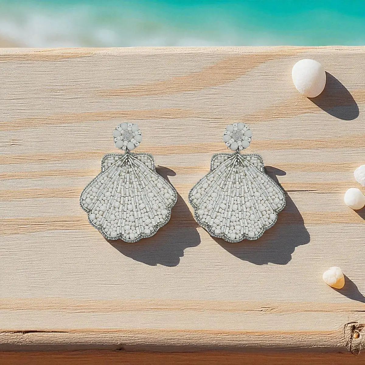 A pair of shell-shaped silver earrings covered in clear crystals, displayed on a light wooden surface with small white shells and a turquoise ocean background.