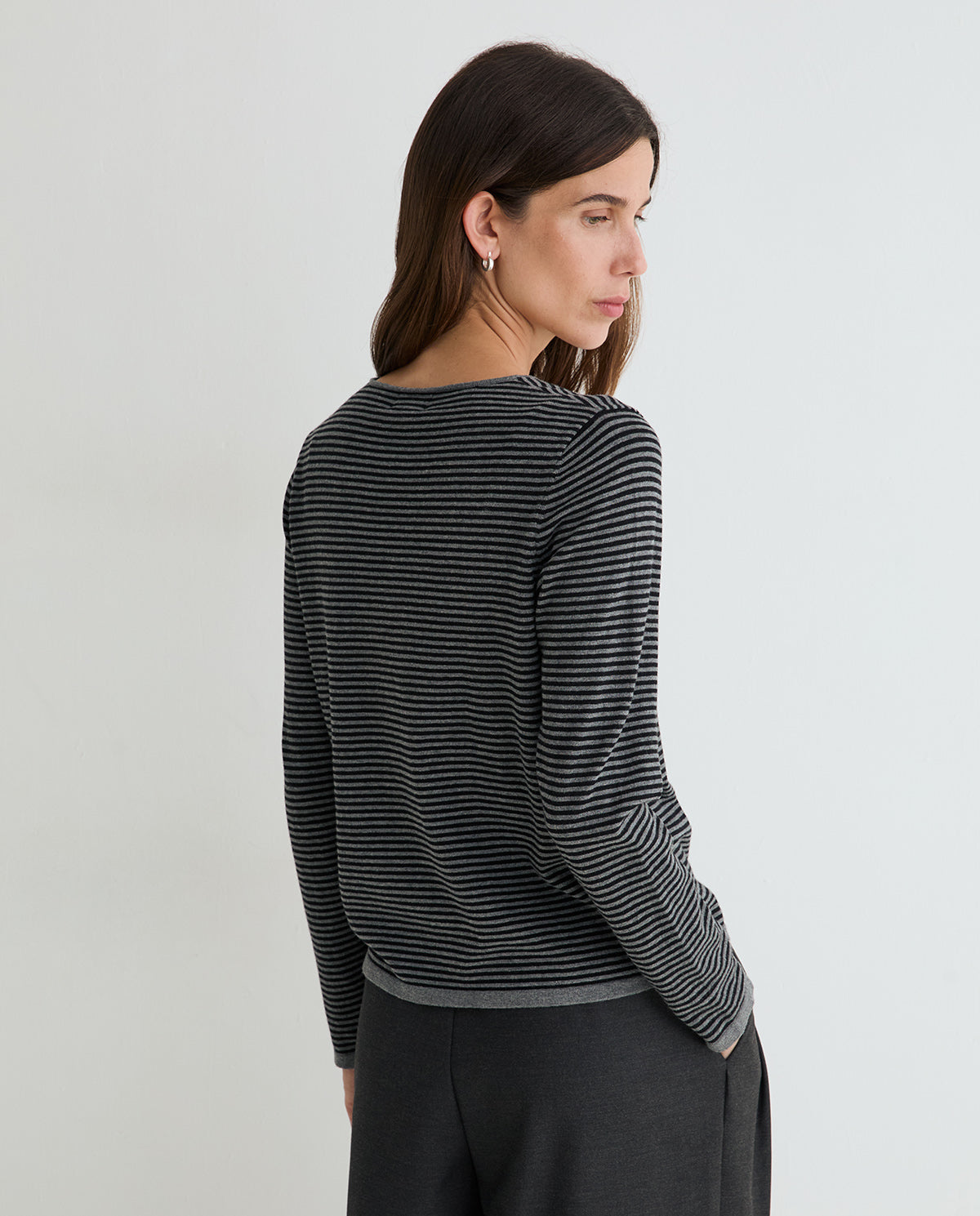 A woman shown from the back wearing a black and gray striped long-sleeve knit top and dark gray trousers, photographed against a plain light gray studio background.