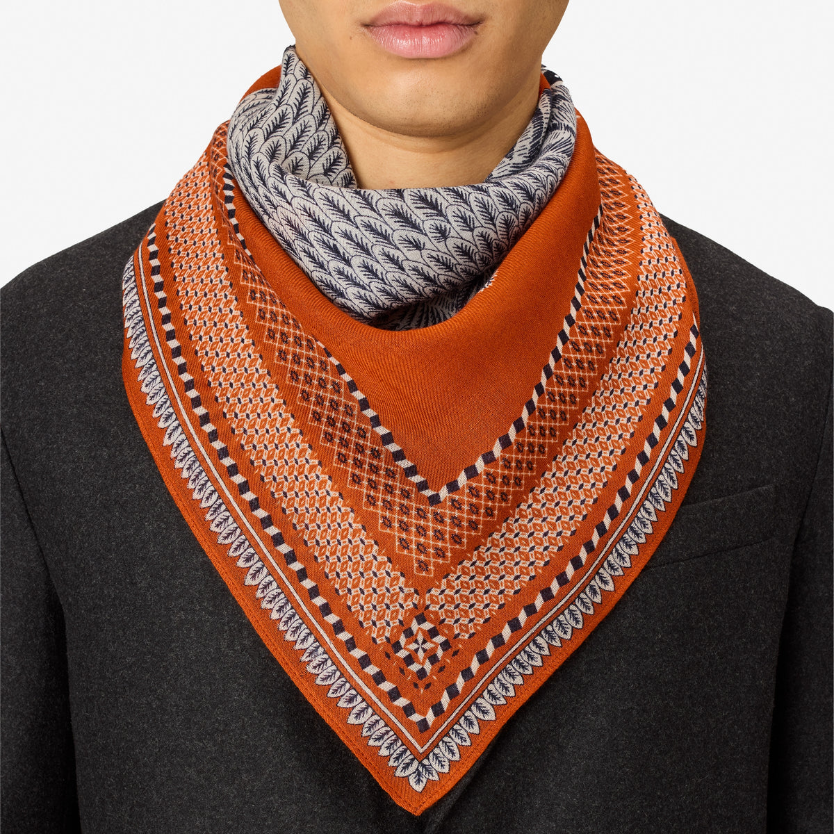 A close-up of a person wearing a patterned orange and gray scarf layered over a gray knit neck warmer, paired with a dark gray coat against a light gray background.