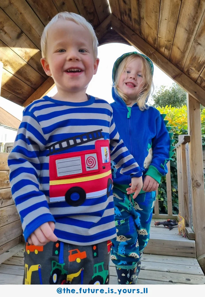 Two children standing on a wooden deck, one wearing a striped shirt with a fire truck design and the other in a blue hoodie.