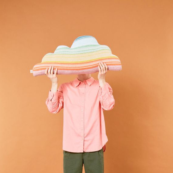 A person wearing a pink button-up shirt, olive green pants, and yellow socks holds a pastel rainbow-striped cloud-shaped pillow in front of their face against a solid warm orange background.
