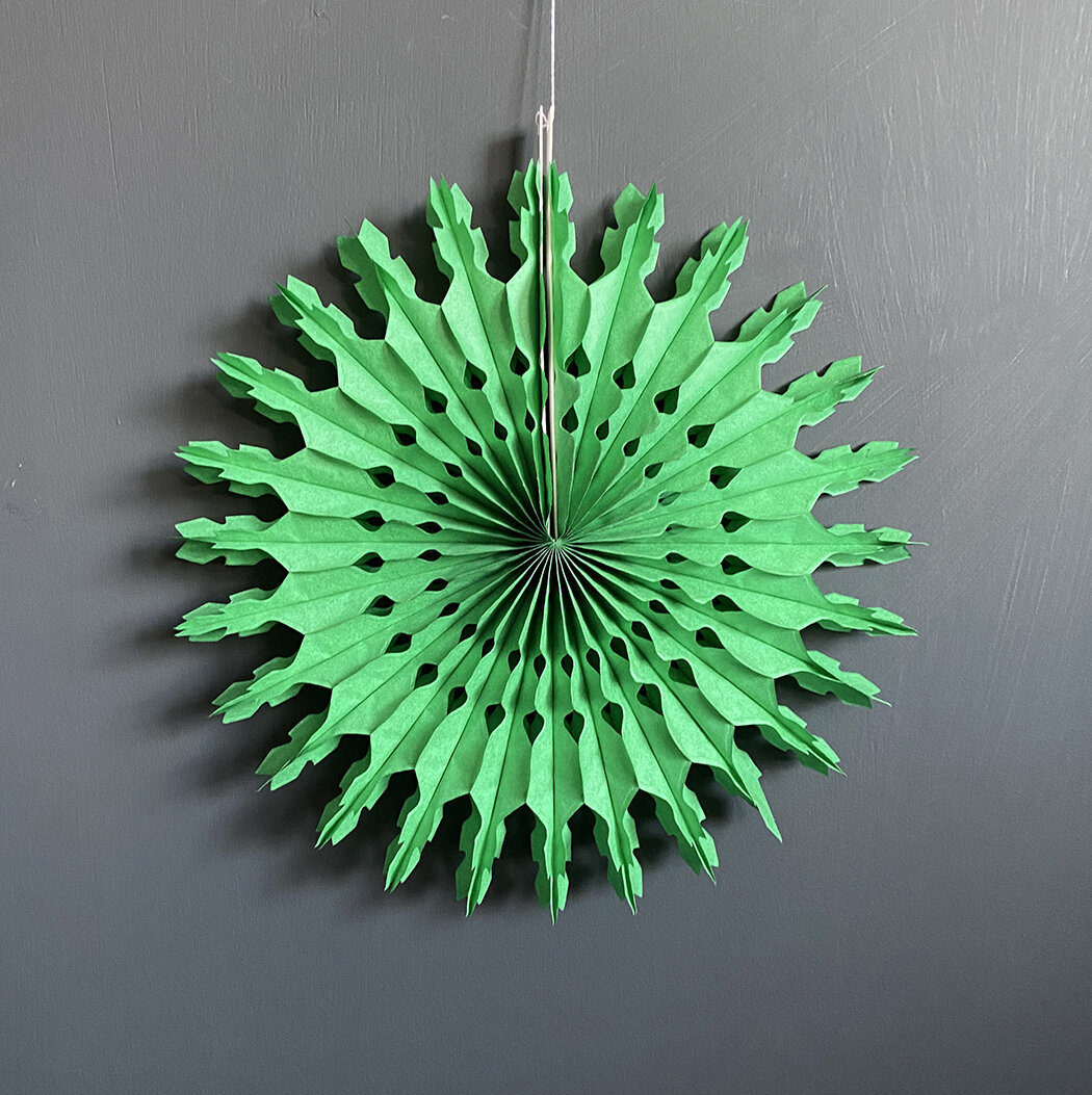Green paper fan decoration with scalloped edges and cut-out details, hanging against a dark gray background.