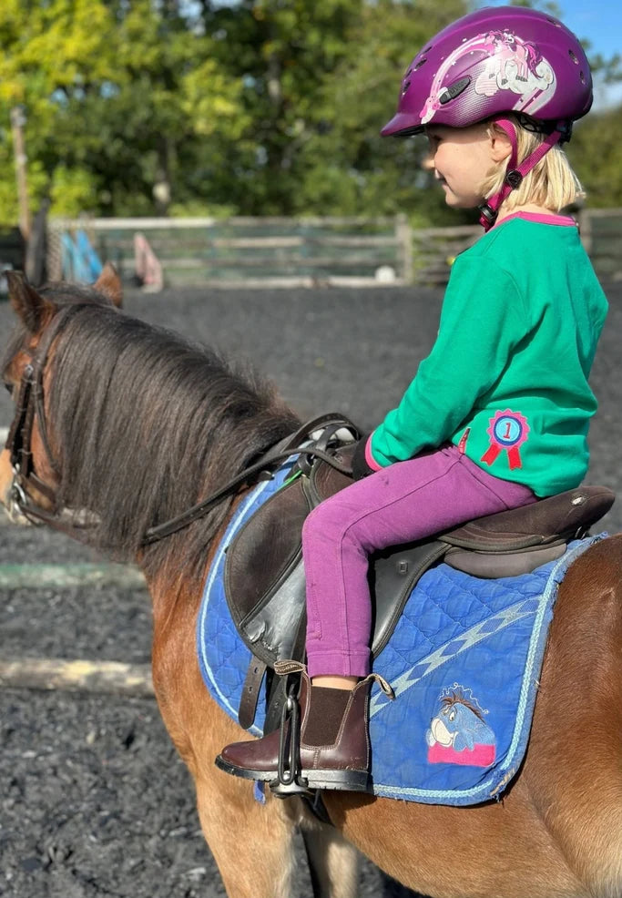 Child riding a horse wearing a helmet and green shirt with a pink ribbon design.