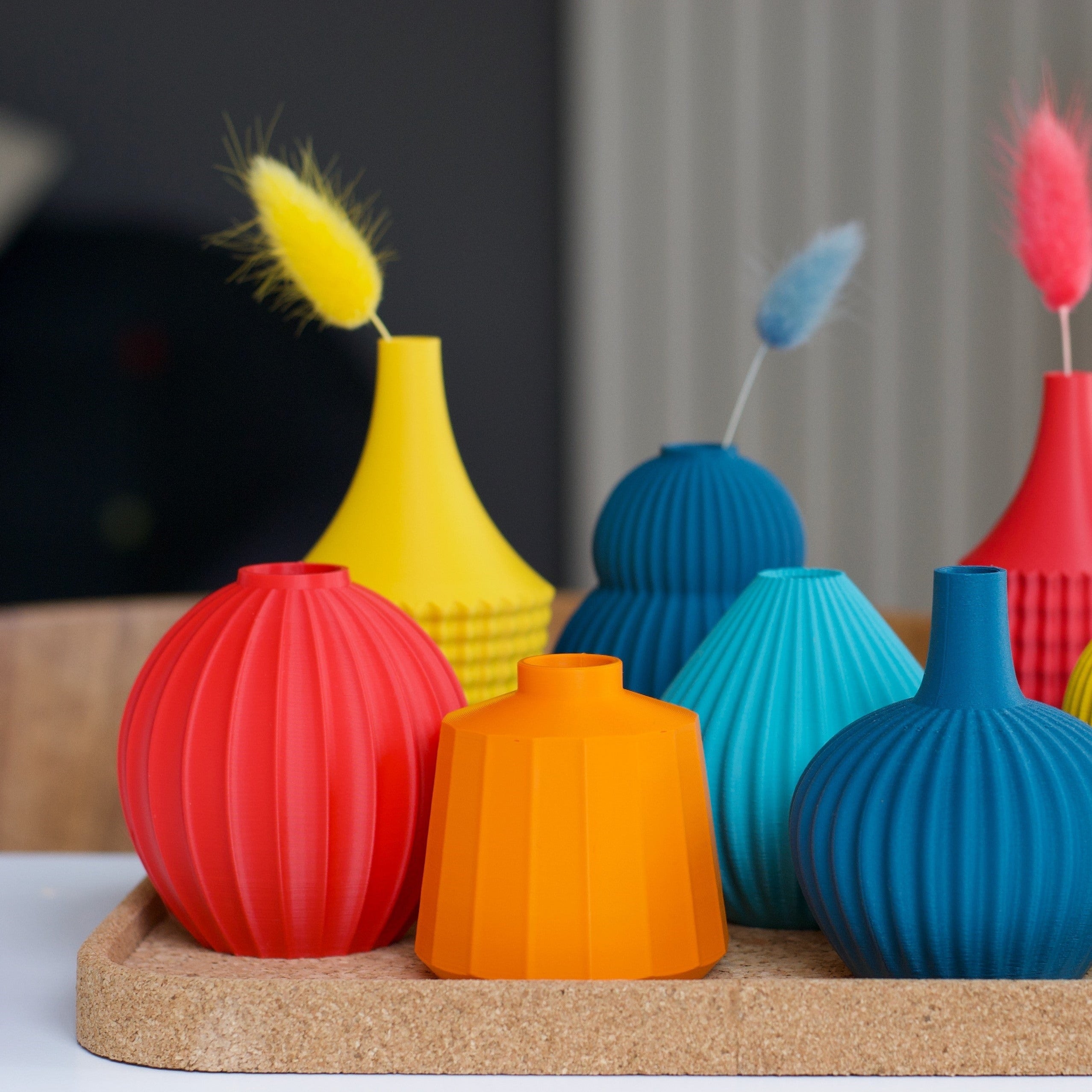 Collection of colorful, ribbed, 3D-printed vases in red, orange, yellow, teal, and blue, displayed on a cork tray.