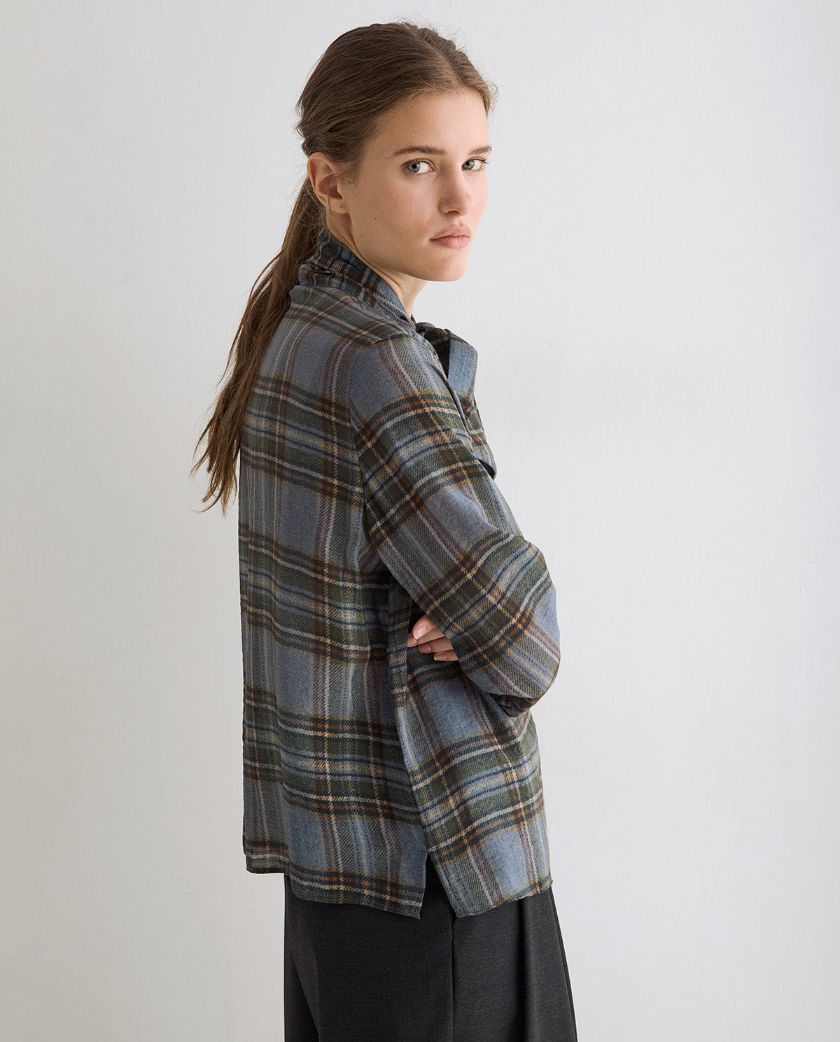 A woman wearing a blue-gray plaid button-up shirt with brown and green stripes, paired with dark gray trousers, photographed against a light gray studio background.