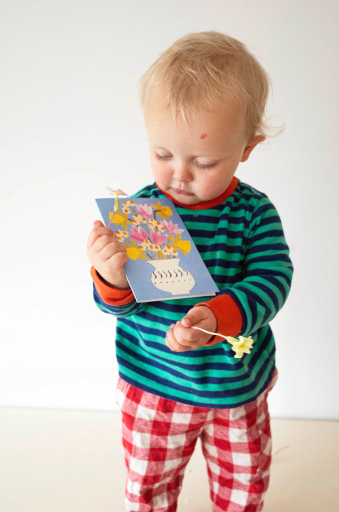 Toddler holding a greeting card depicting a floral arrangement in a vase.  The card is light blue with colorful flowers.
