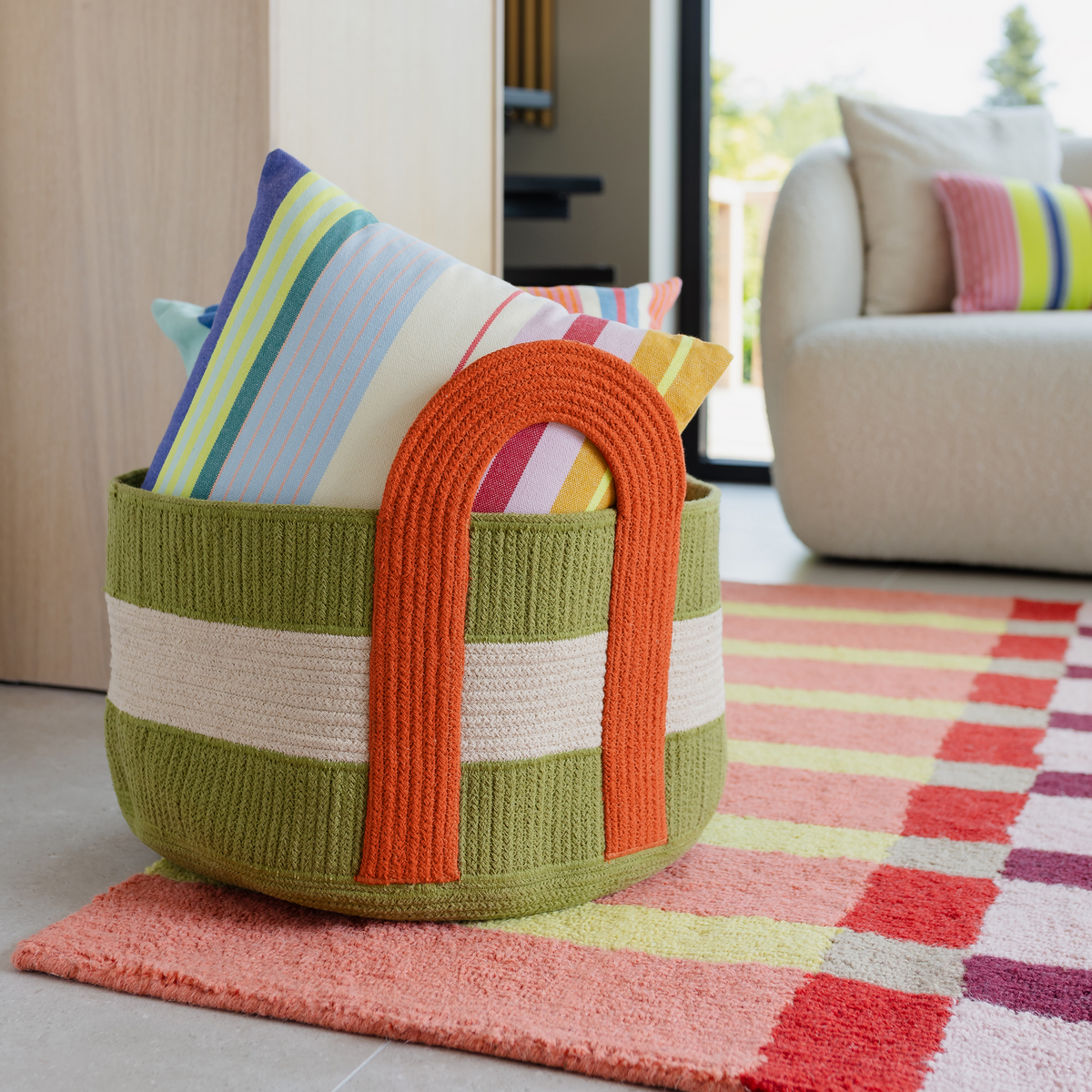 A woven storage basket with a green and white striped body and bright orange handles, filled with colorful striped throw pillows, placed on a pink and red checkered rug in a modern living room setting.