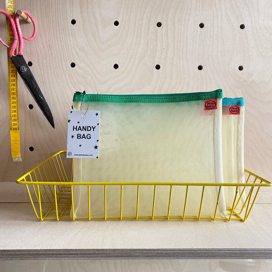 Two pale yellow mesh zipper pouches with green and teal zipper trim, in a yellow wire basket. A tag labels them "Handy Bag".