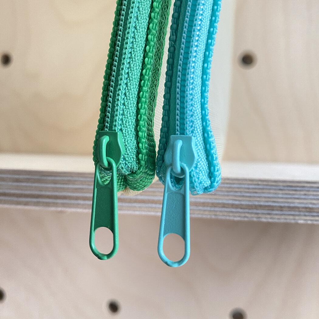 Two small zippered pouches; one teal, one green. Zips are pastel-colored plastic.
