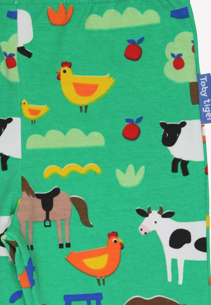 A close-up of a children's garment featuring a colorful cartoon farm print on a bright green background. The pattern includes yellow and orange chickens, a brown horse, a black-and-white cow, red apples, and light green clouds.