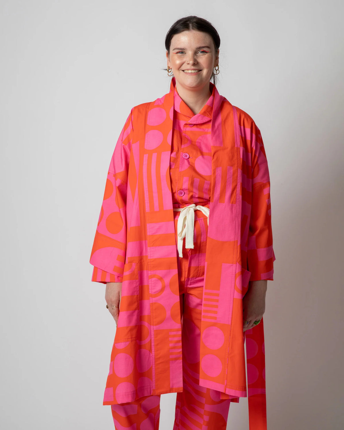 A woman modeling a matching two-piece outfit with a vibrant abstract geometric pattern in orange and pink, featuring a long open-front robe-style top and coordinating pants with a cream tie belt.