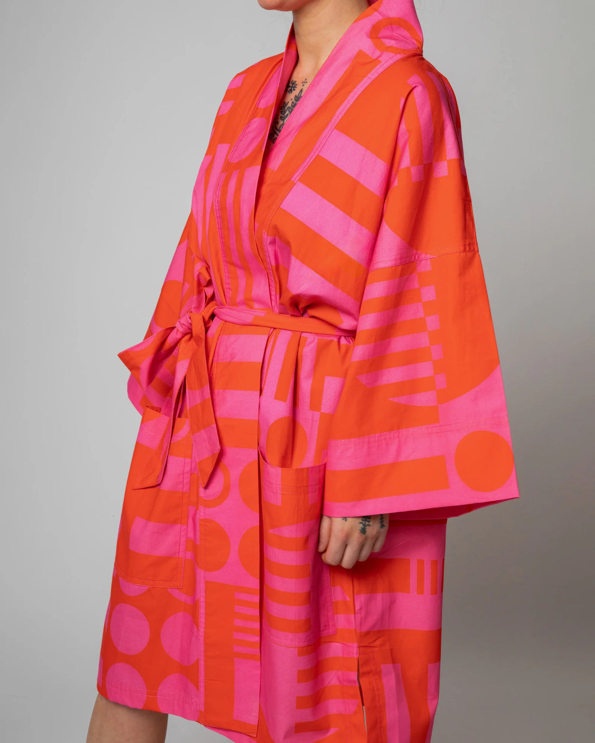 A person wearing a kimono-style robe with a bold geometric pattern in bright orange and pink, photographed against a plain light gray studio background.