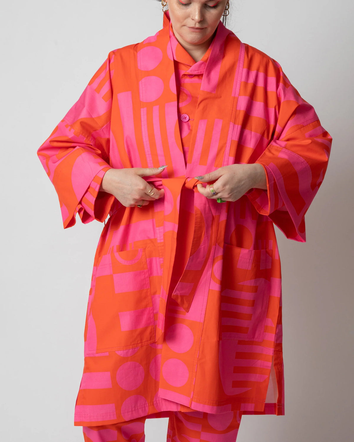 A model wearing a vibrant orange and pink geometric print kimono-style robe with a matching tie belt, shown against a plain light gray studio background.