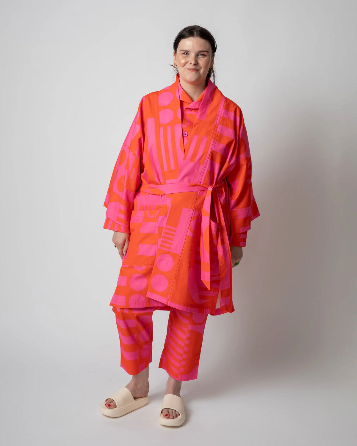 A model stands against a light gray studio background wearing a bright pink and orange kimono-style robe with a matching patterned pair of pants and cream slide sandals.