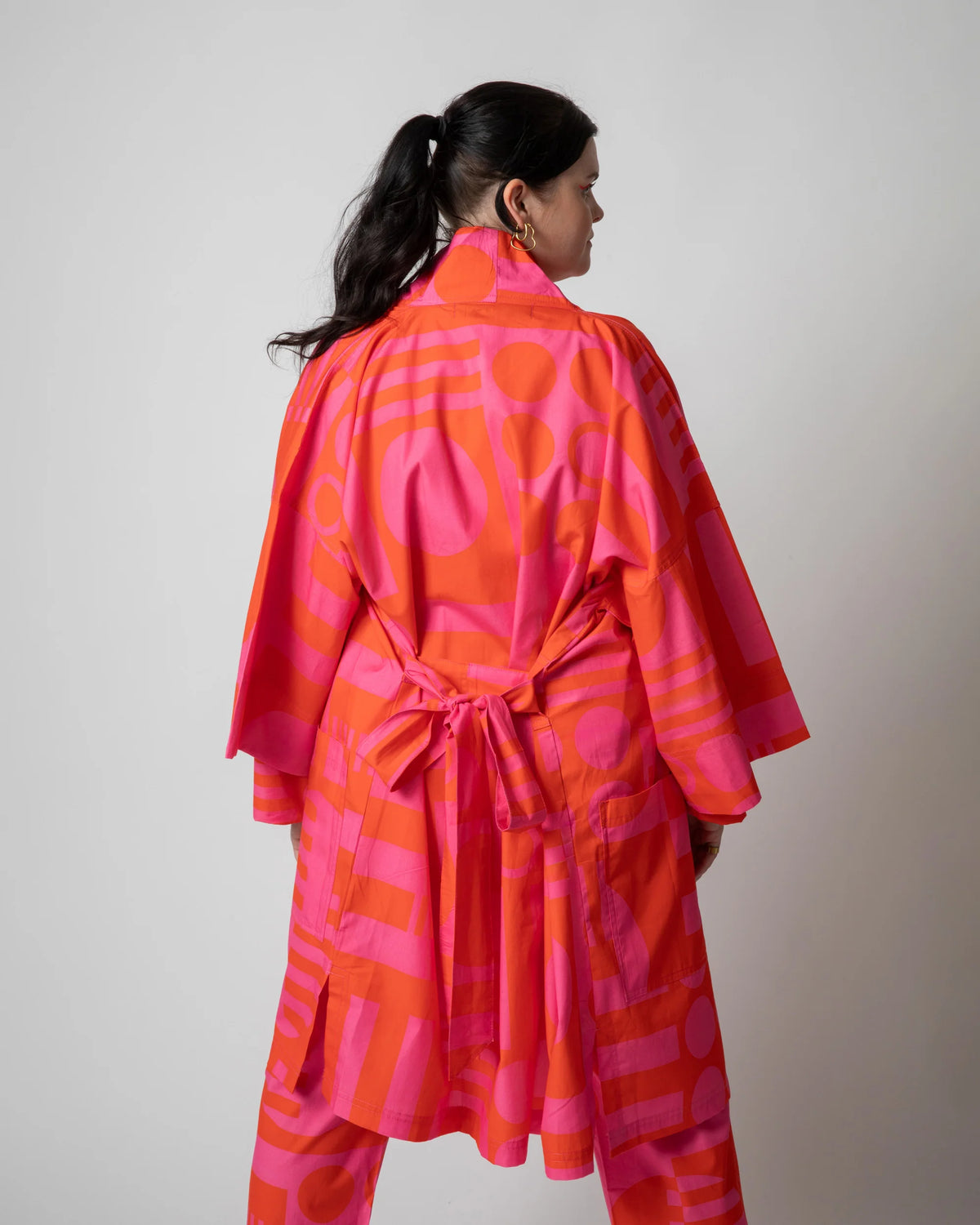 A woman shown from the back wearing a bright pink and orange patterned kimono-style robe with a tie belt, photographed against a plain light gray studio background.