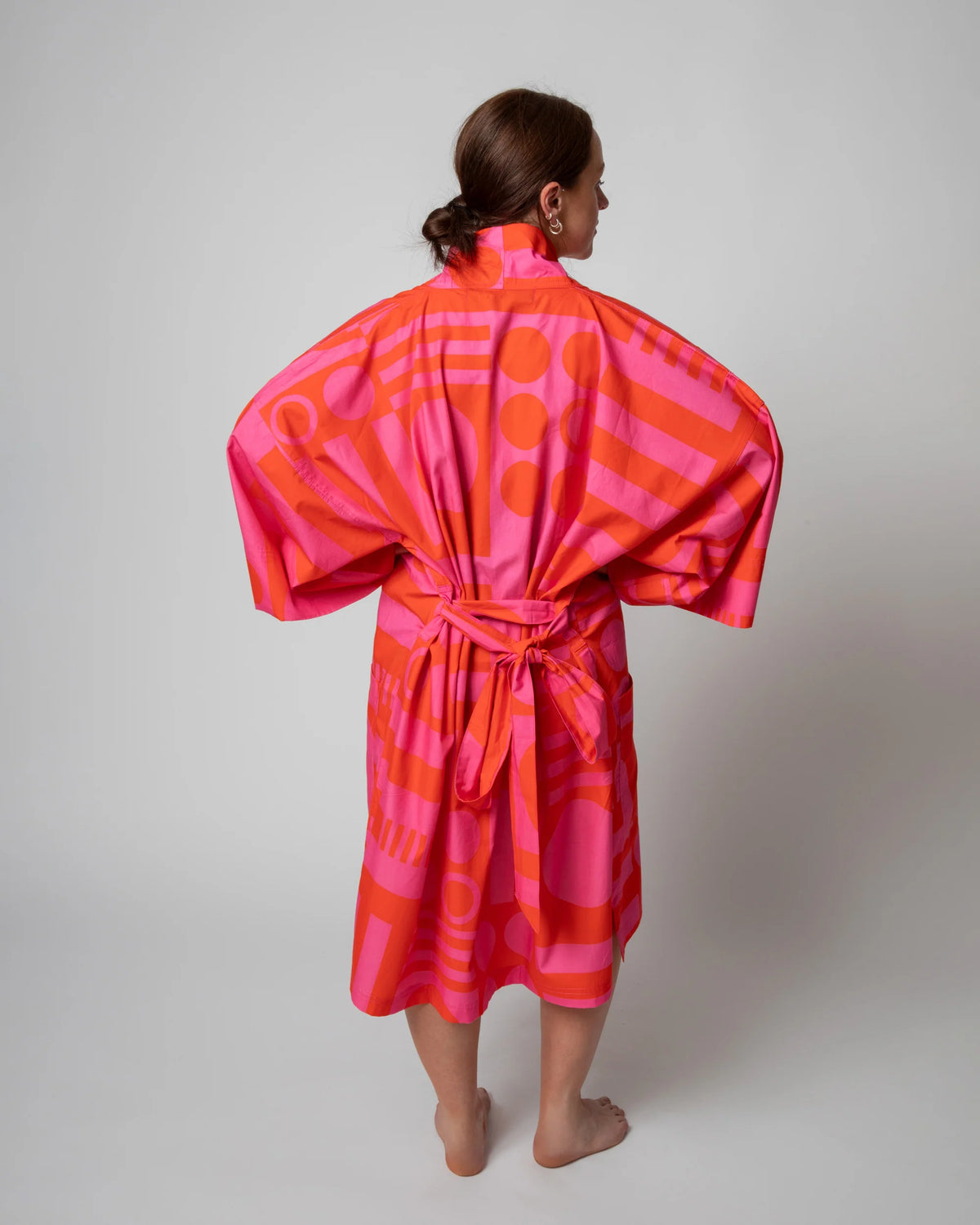 A woman stands barefoot against a light gray studio background, viewed from the back, wearing a loose, high-neck robe or kimono-style garment in bright pink and orange with an abstract geometric print and a tied waist belt.