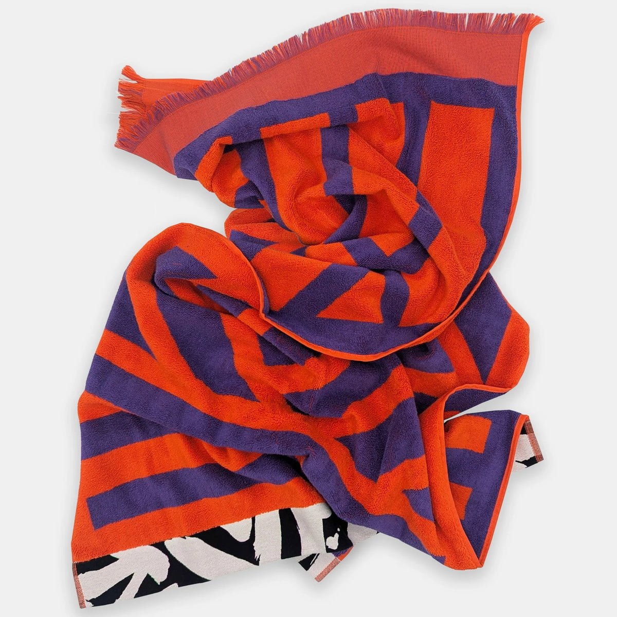 A lightweight woven scarf with a bold geometric pattern in bright orange and deep purple, featuring fringed edges and a black and white patterned reverse side, displayed on a light gray background.