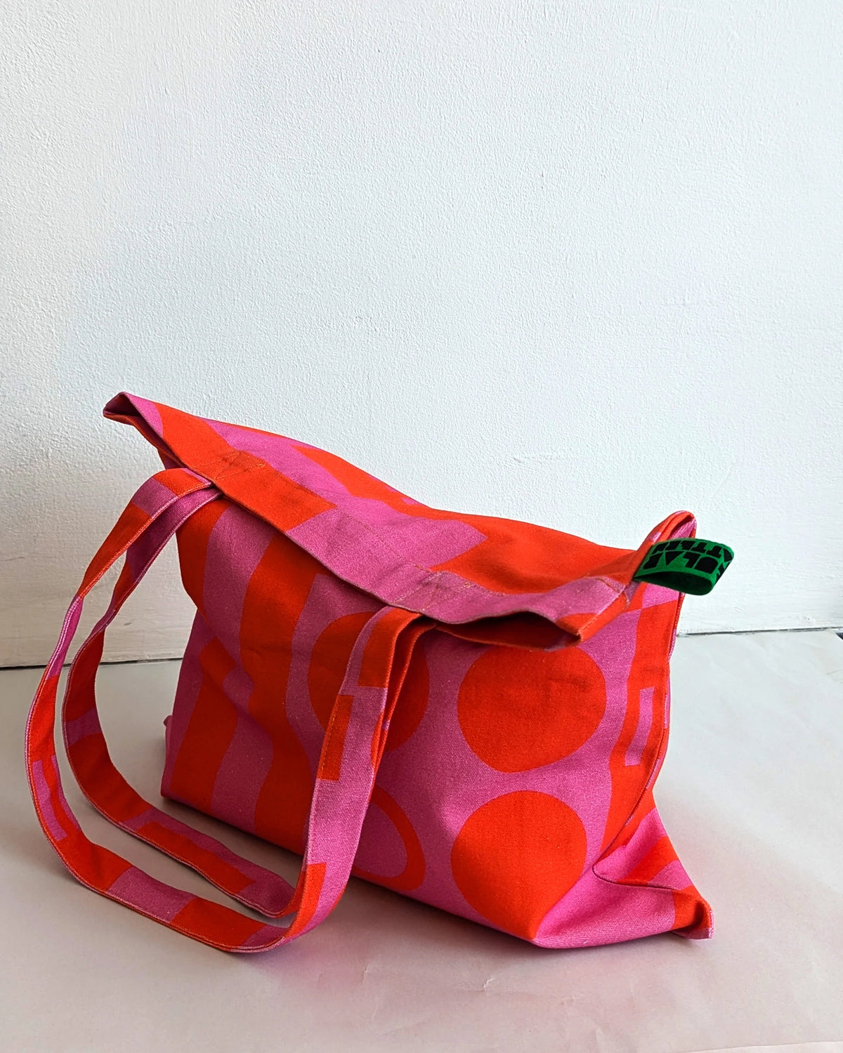 A fabric tote bag with a bold geometric pattern in vibrant pink and orange-red, featuring a matching shoulder strap and a small green fabric tag.