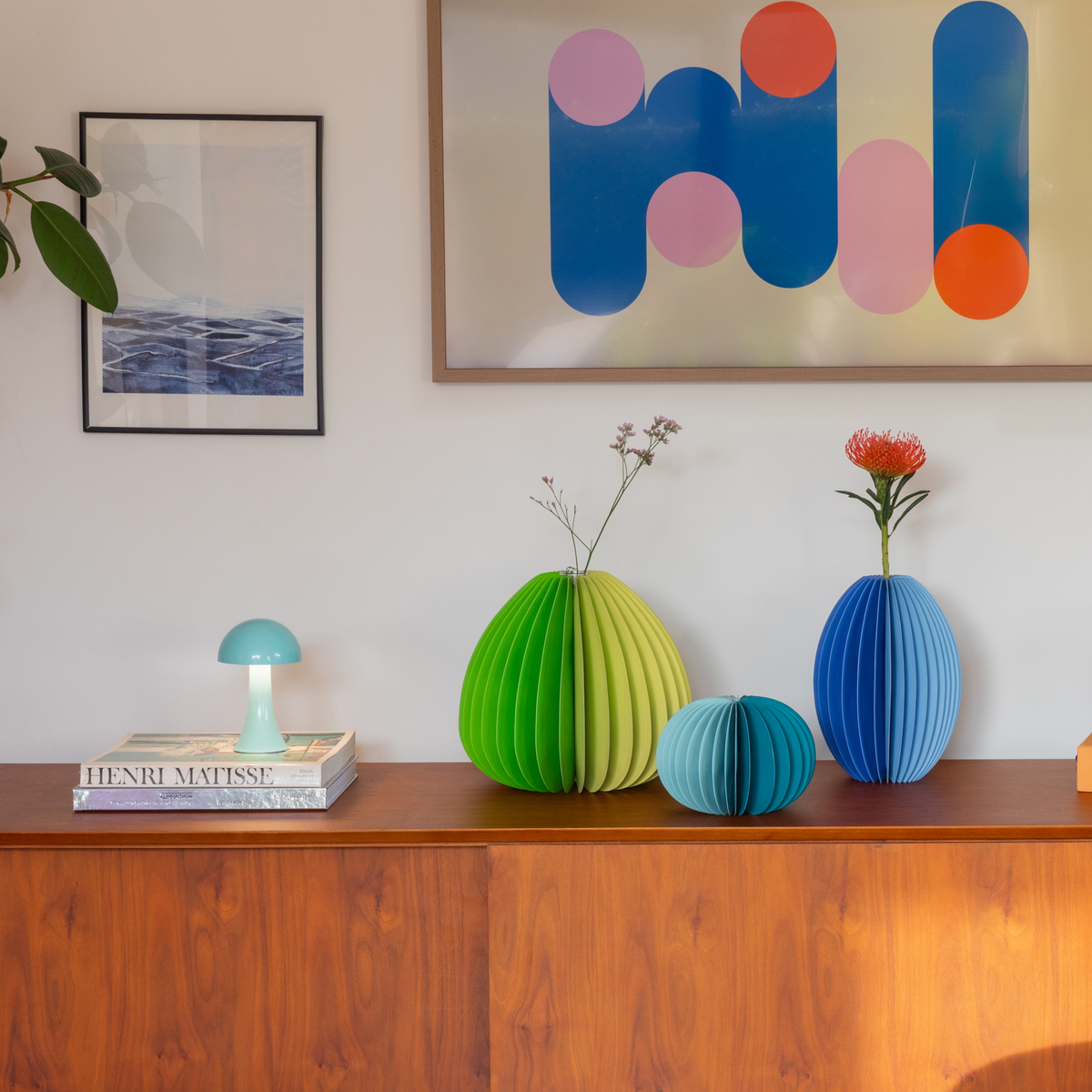 Colorful paper vases and a lamp on a wooden surface with abstract art in the background.