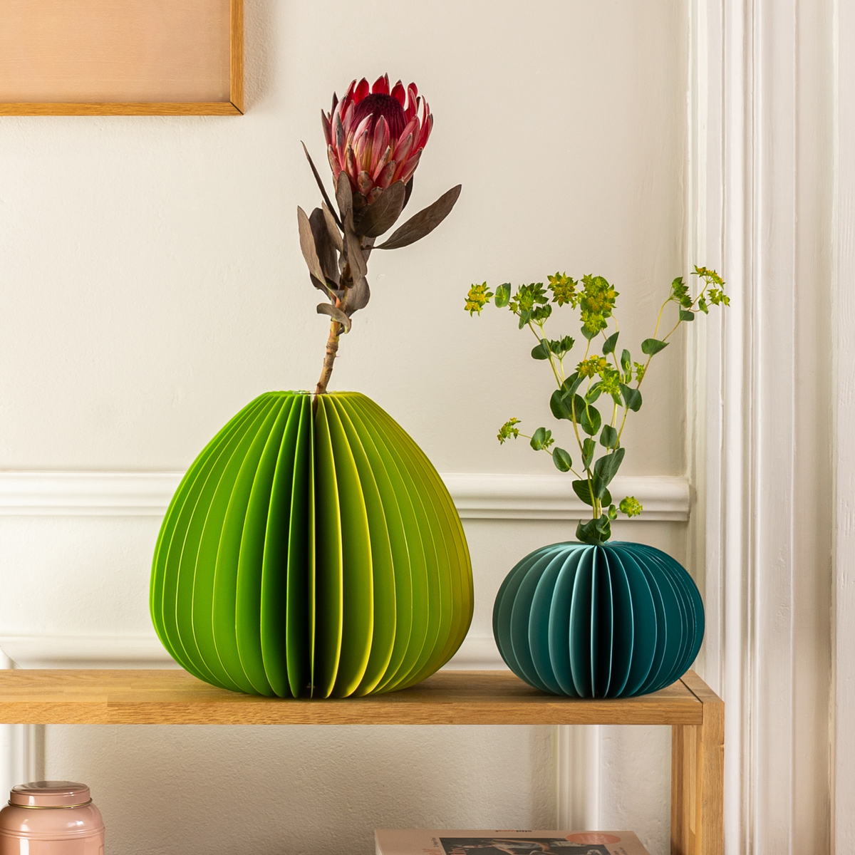 Two colorful paper vases with plants on a wooden shelf against a light wall.
