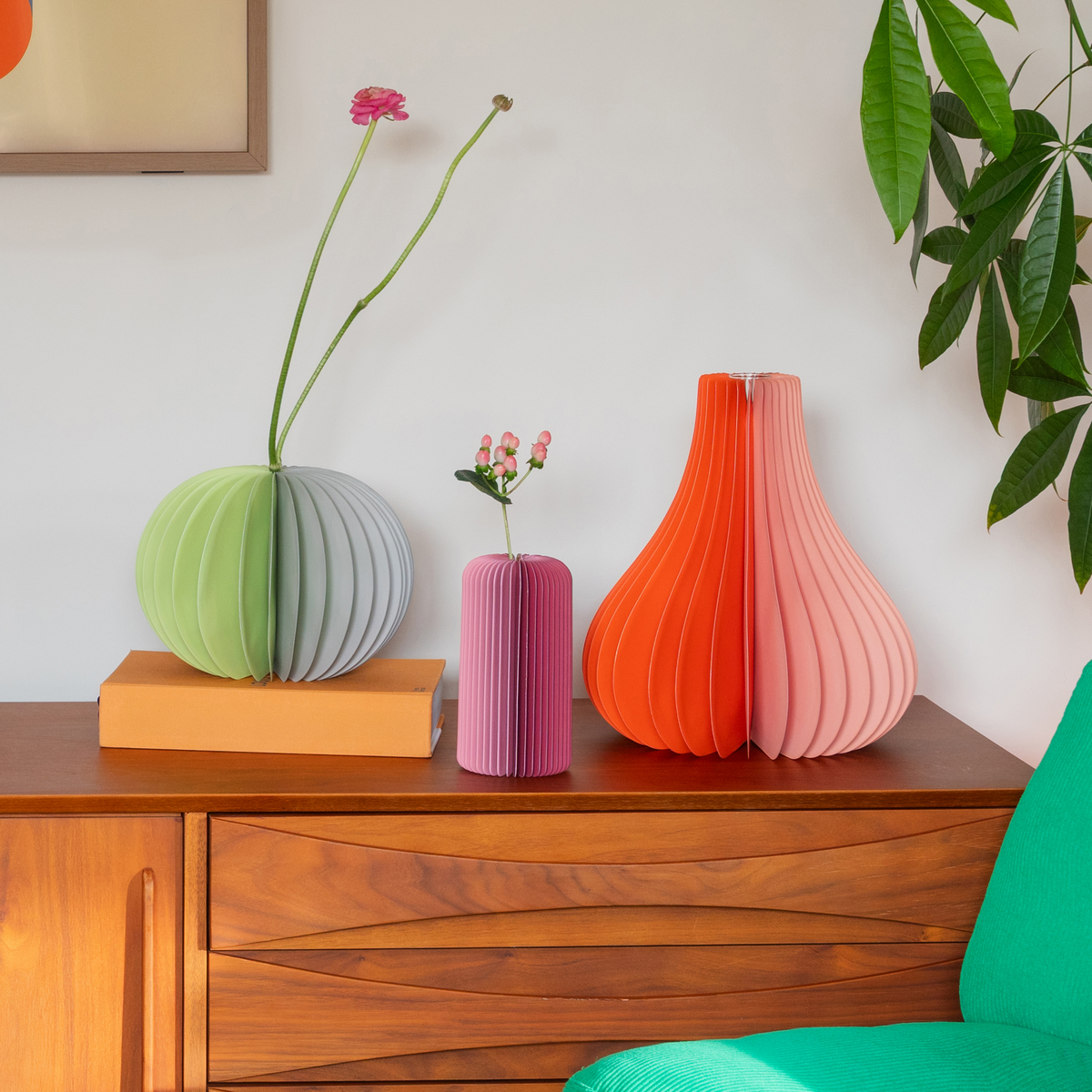 Three modern accordion-style paper vases in green, pink, orange, and coral-pink, displayed on a wooden sideboard with flowers in a styled interior setting.