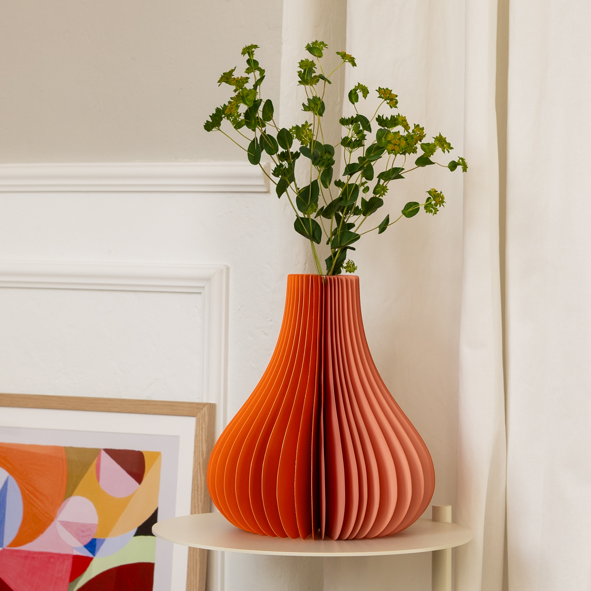 Orange paper vase with greenery on a white surface, with a colorful abstract painting in the background.