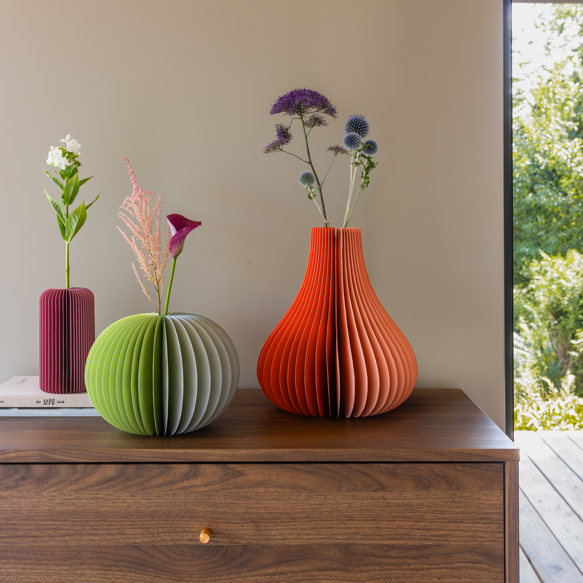 Three sculptural paper fiber vases in green, orange, and magenta displayed on a dark wood sideboard, with dried and live flowers inserted in the vases, set against a neutral beige wall with a window view of green foliage on the right.