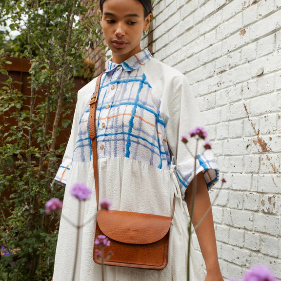 Brown leather crossbody bag with a flap closure, worn by a model in a white and blue patterned dress.