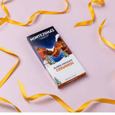A box of Montezuma's Blonde Chocolate Cinnamon chocolate bar, styled on a light pink background with flowing yellow ribbons.