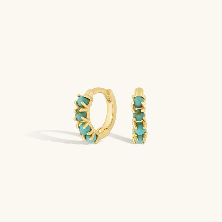 A pair of gold huggie hoop earrings with small turquoise-colored stones set along the front, displayed on a clean off-white background.