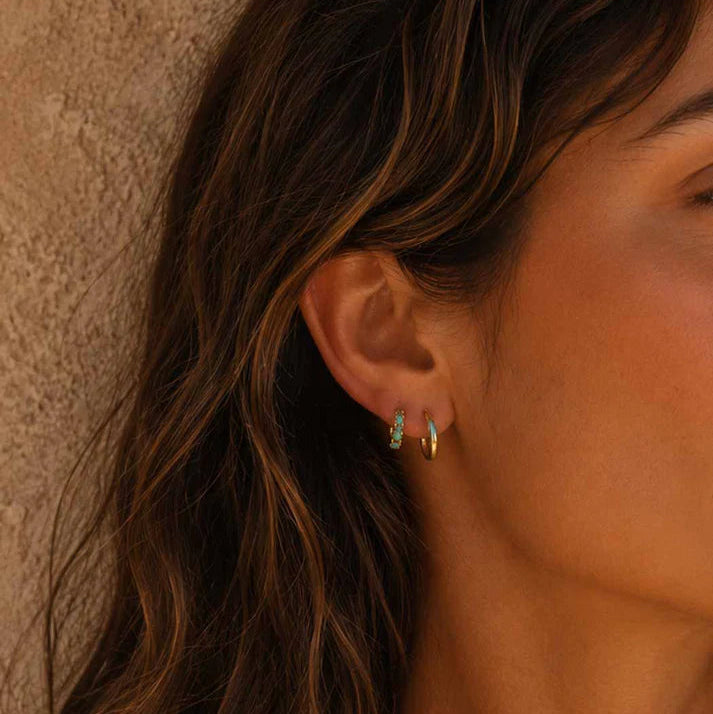 A close-up side profile of a woman with long wavy brown hair wearing a pair of small gold hoop earrings with tiny light blue stone accents, set against a textured beige wall.