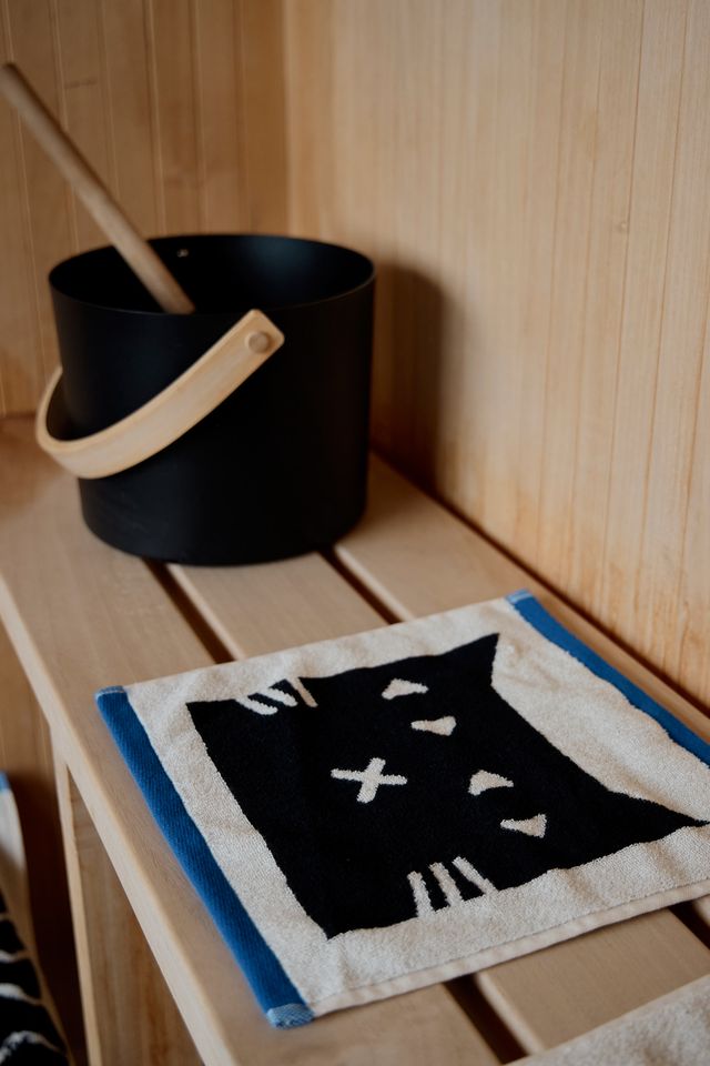 A close-up of a woven textile piece featuring a black cat face motif with white details, bordered in blue and beige, resting on a light wood slatted surface next to a black bucket with a wooden handle.