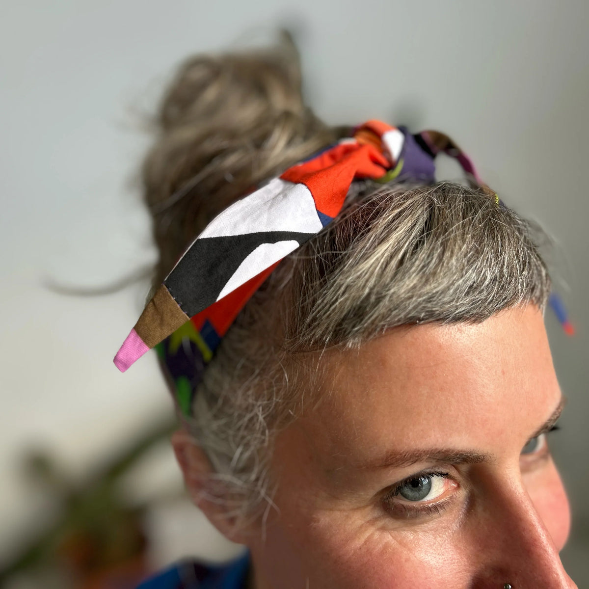 A close-up lifestyle shot of a woman with gray hair in a messy bun wearing a colorful, patterned fabric headband. The headband features a geometric patchwork design with red, white, black, purple, and brown sections.